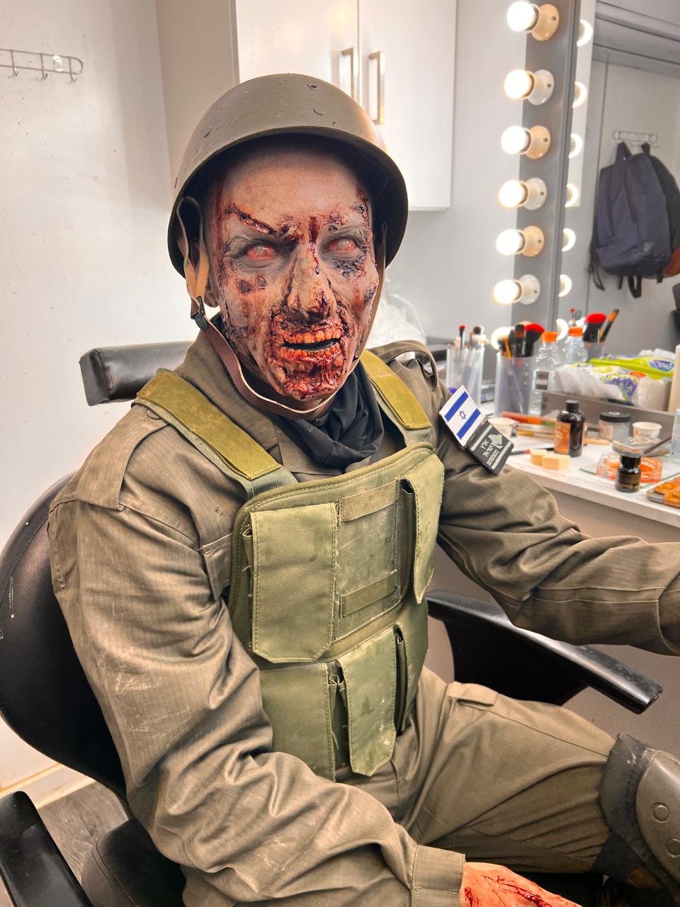 Frontal seated helmeted character in a makeup room with extensive red/dark facial injury-like texture and light red eye appearance; mirror bulbs, products and costume are visible.