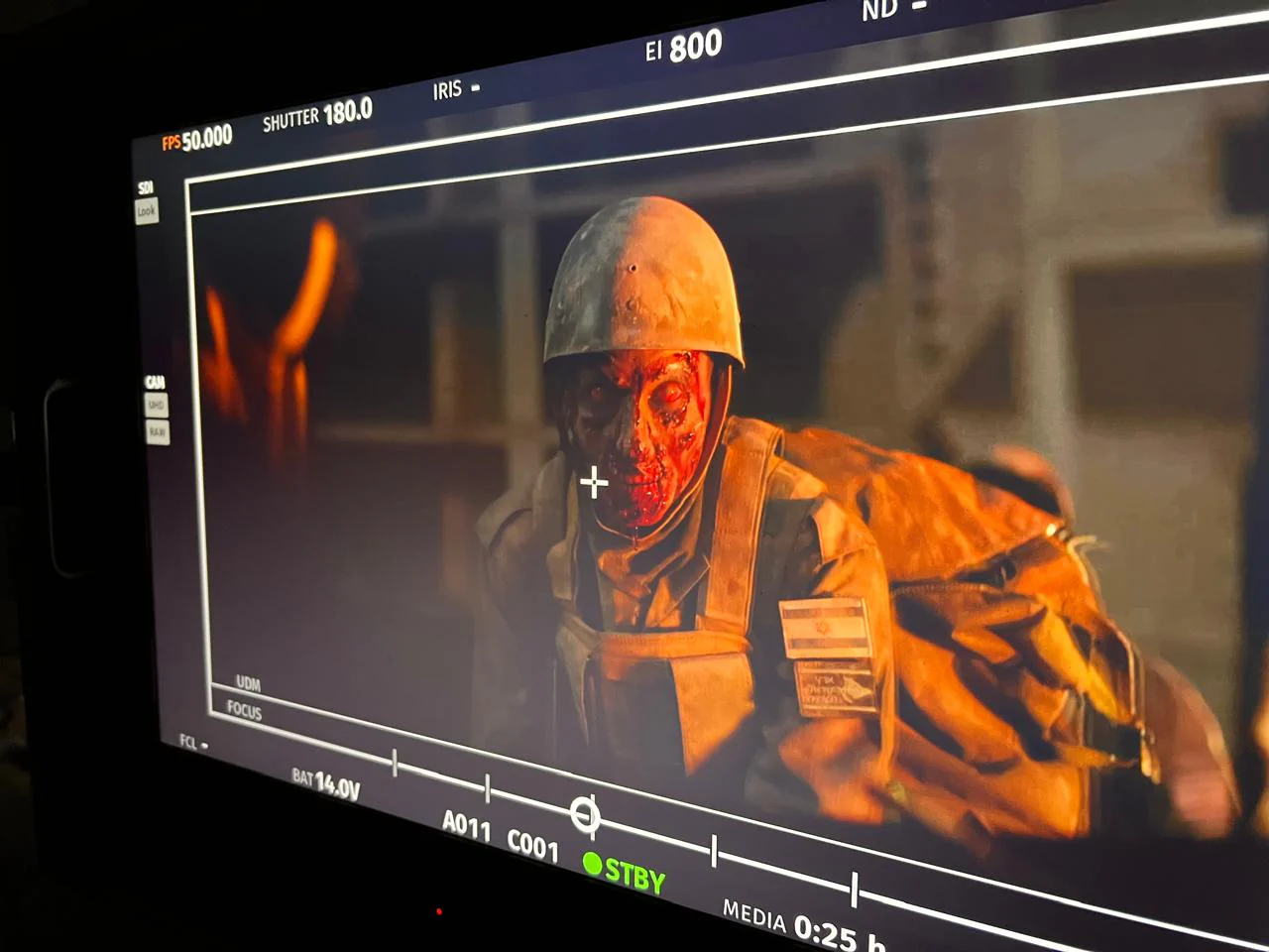 Angled production-monitor photograph of a helmeted character with red/dark facial detailing under warm orange scene light; display settings and framing marks remain visible.