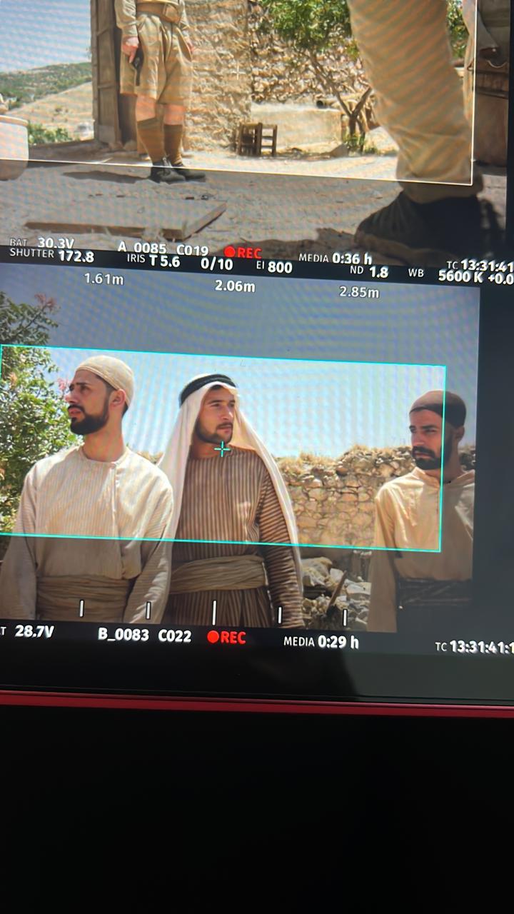 Portrait photo of two stacked monitor views: upper courtyard/equipment detail, lower view of three bearded performers in pale period-style clothing and head coverings beside a stone wall.