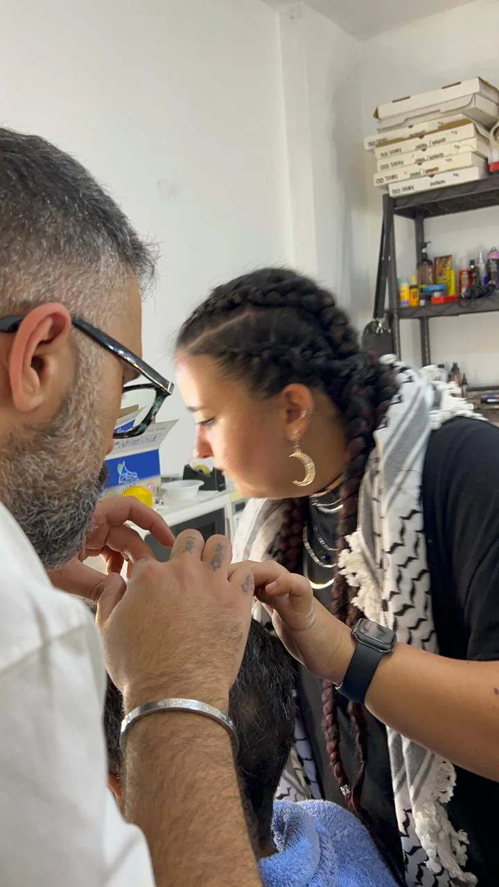 Side BTS photograph of a braided-hair person working near a seated bearded performer's lower face in a room with product shelves; a second person and towel are partly visible.