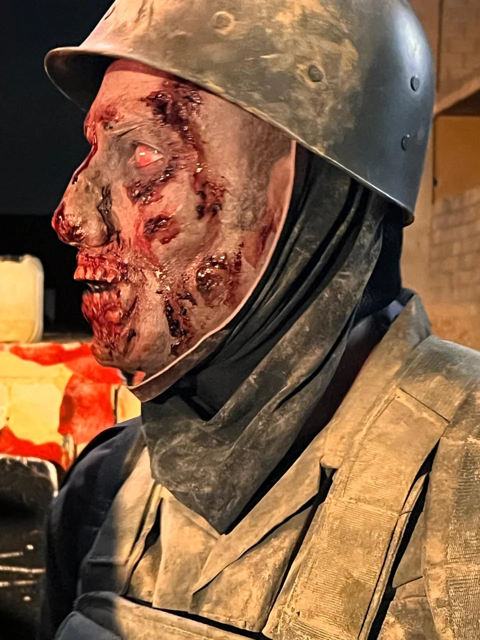 Close side-profile character photograph with helmet, neck covering and heavily textured red/dark facial injury-like details including the eye, cheek and mouth.