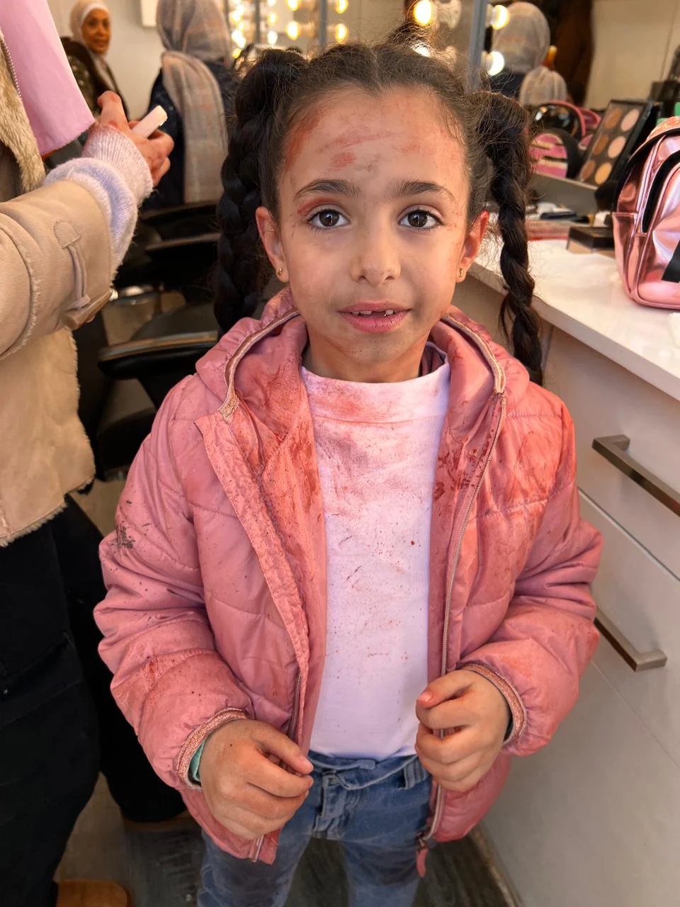 Child performer in a makeup-room setting, braided hair, pink jacket and pale shirt with dirt/red staining; subtle red forehead marks and scattered tonal detail on face and hands.