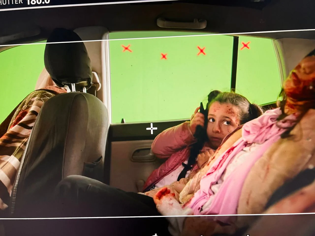 Photographed production-monitor scene inside a vehicle: a child in pink clothing holding a phone, red forehead detailing, adults partly visible and green-screen tracking marks outside the windows.