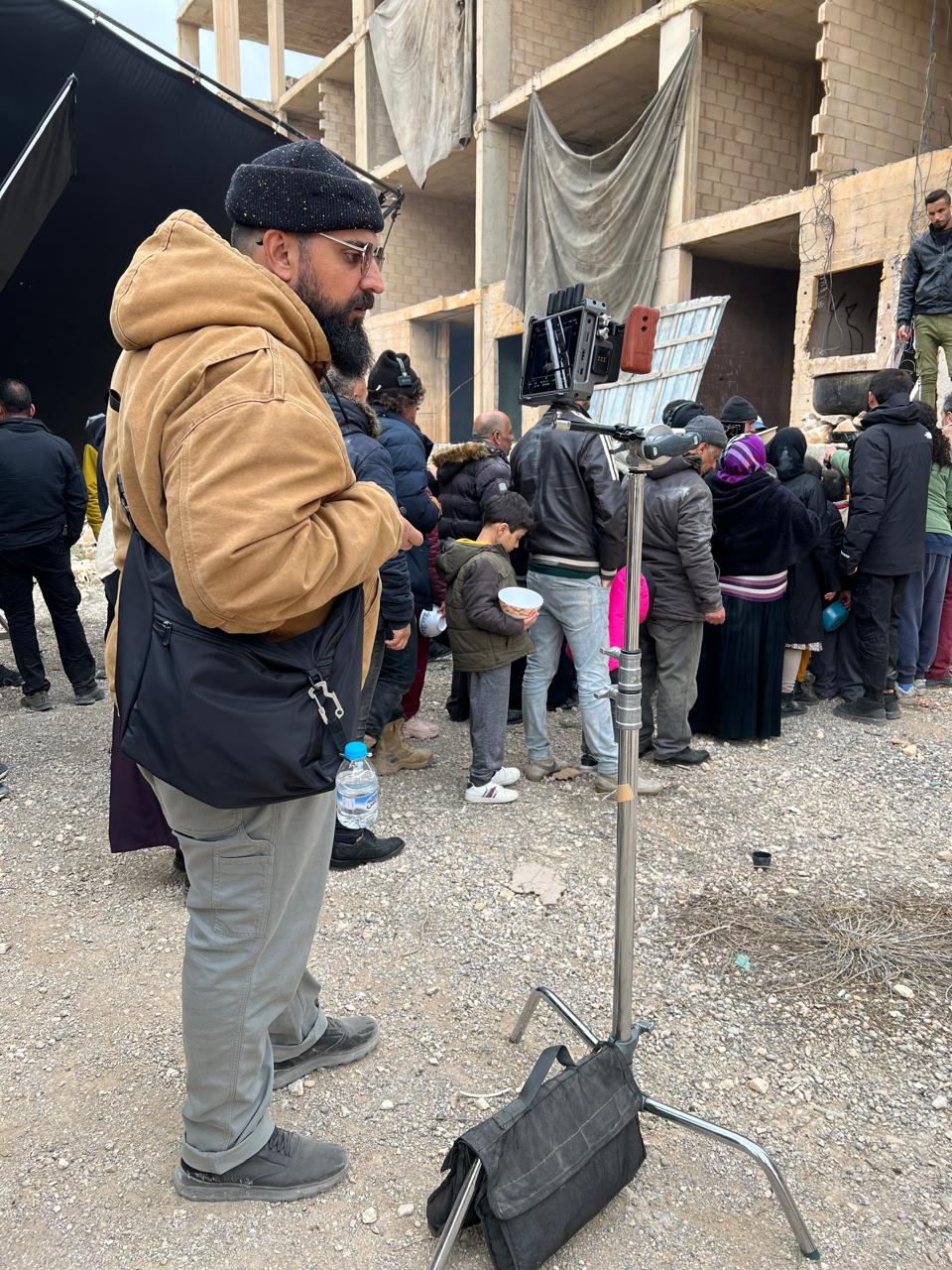 Behind-the-scenes set photograph with a person beside a production monitor, a large group of people and unfinished/damaged-looking masonry, hanging fabrics and filming equipment. No clear makeup application or close makeup detail is shown.
