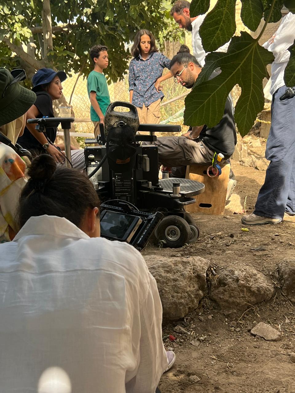 Outdoor filming BTS photograph with a large camera rig, monitoring equipment and several people. Makeup application is not visible.