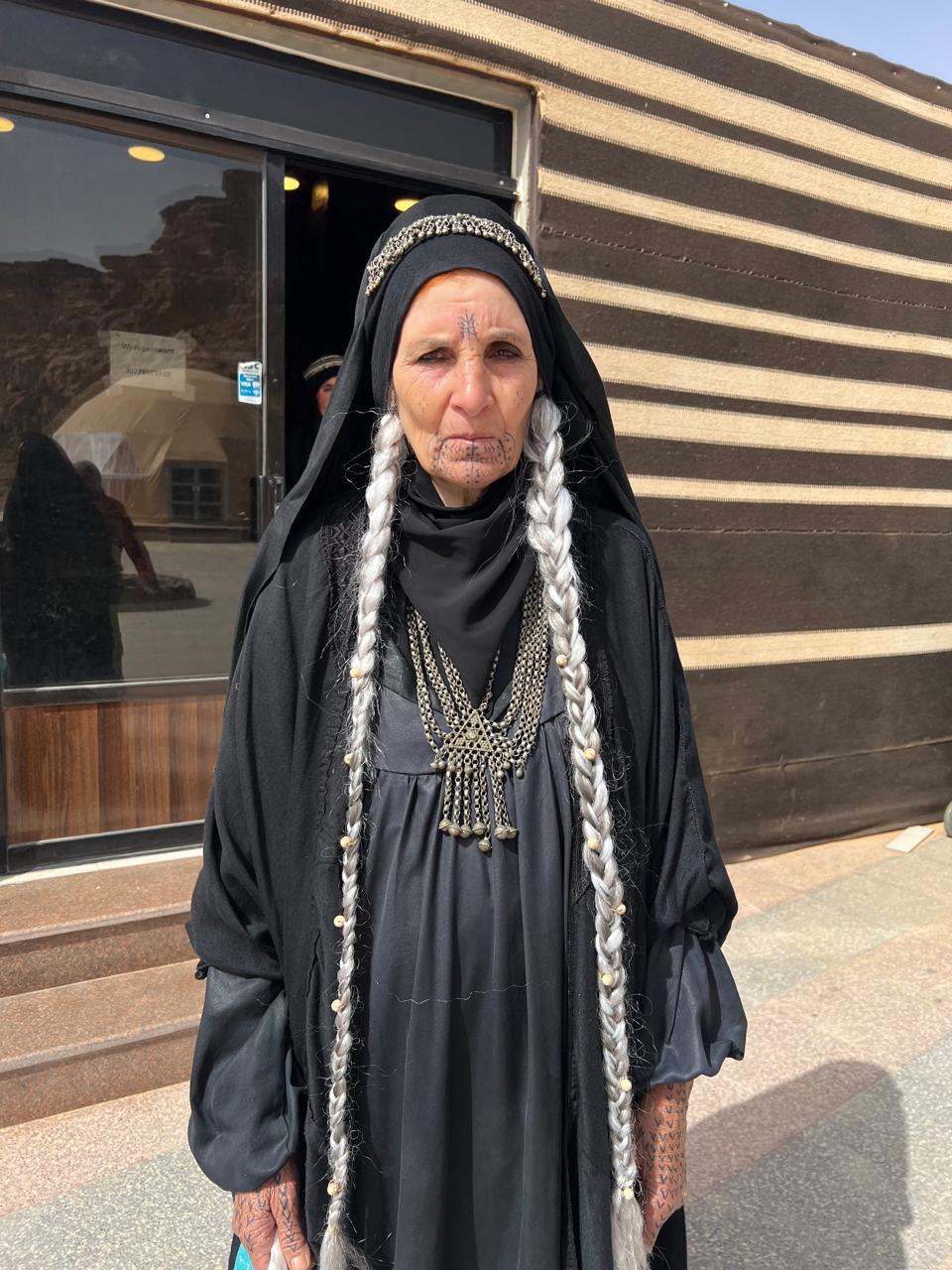 Outdoor character portrait in black clothing/head covering with long gray braids, silver head adornment and necklaces, drawn facial and hand motif-like details.