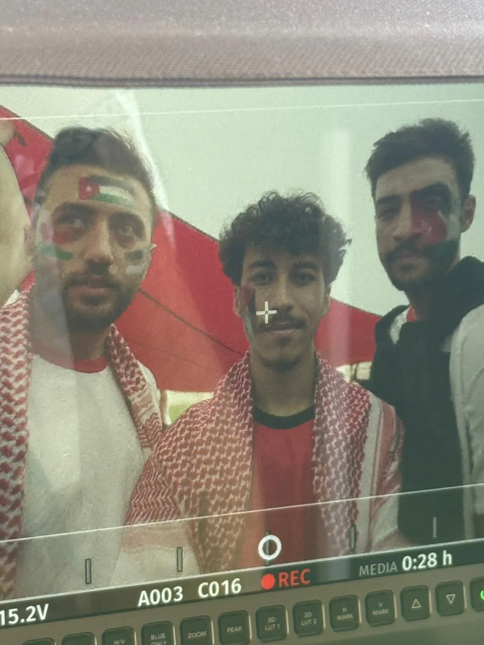 Production-monitor photograph showing three performers in a group with flag-style face paint and patterned scarves, large red/green/white background flag; display crosshair/settings and reflection remain visible.