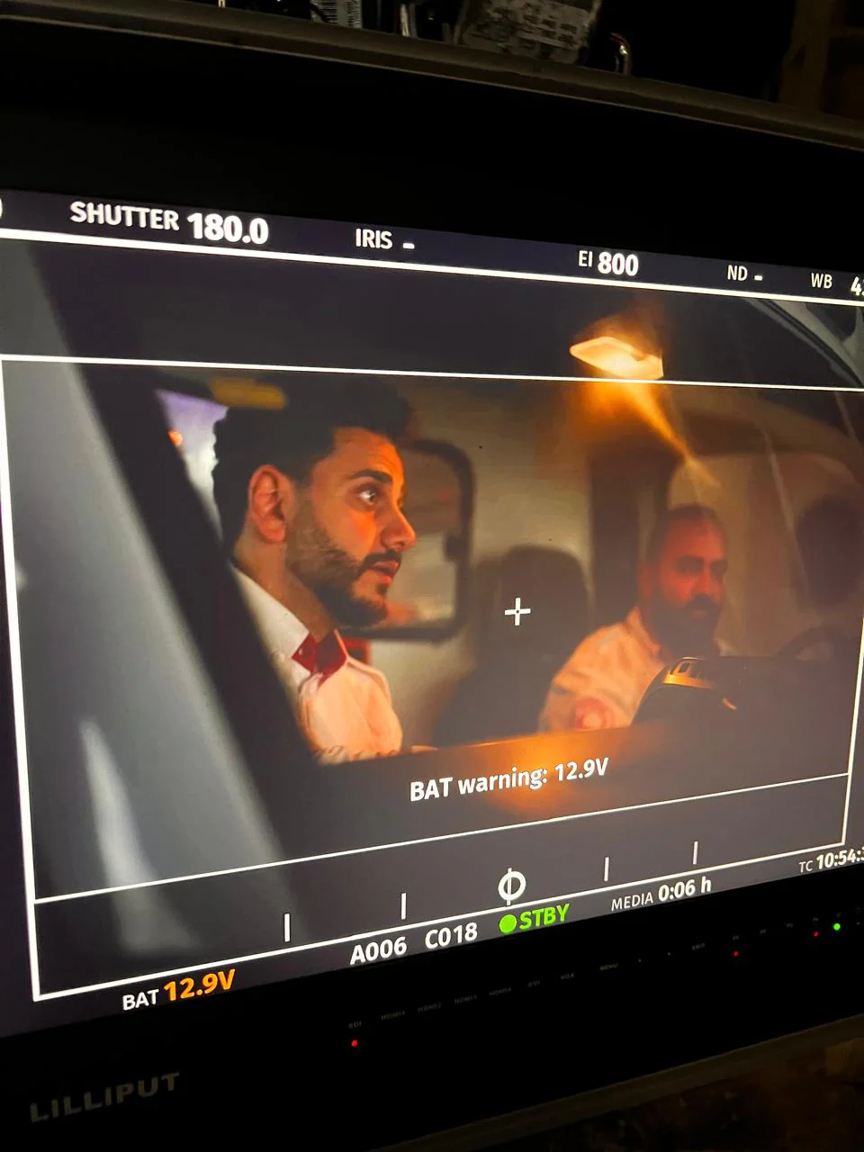 Angled monitor photograph showing two bearded performers in pale shirts inside a vehicle under warm lighting; display overlays and battery warning remain in the original.