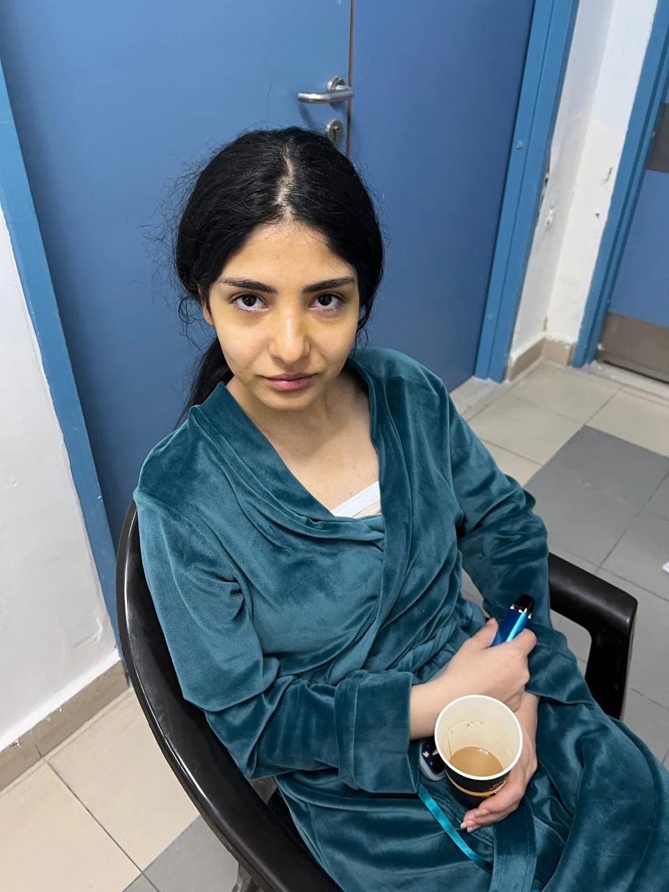 Seated performer wrapped in a teal cloth in a blue-door room, face showing pale/yellow-green tonal treatment and darker under-eye shading; cup and blue item held in hands.