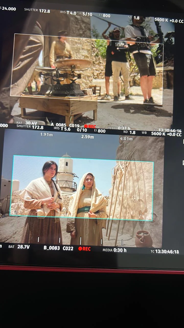 Portrait photo of two stacked production-monitor views of a stone courtyard: upper view includes equipment/crew, lower view shows two performers in pale period-style clothing and head coverings.