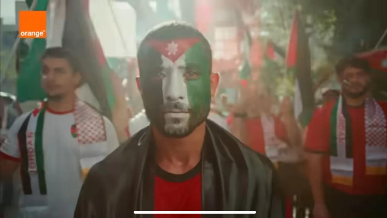 Landscape supplied scene frame with Orange logo at upper left: central performer with face painted in black/white/green/red flag-style shapes and white star, other performers and flags behind; white phone home-indicator bar at bottom.