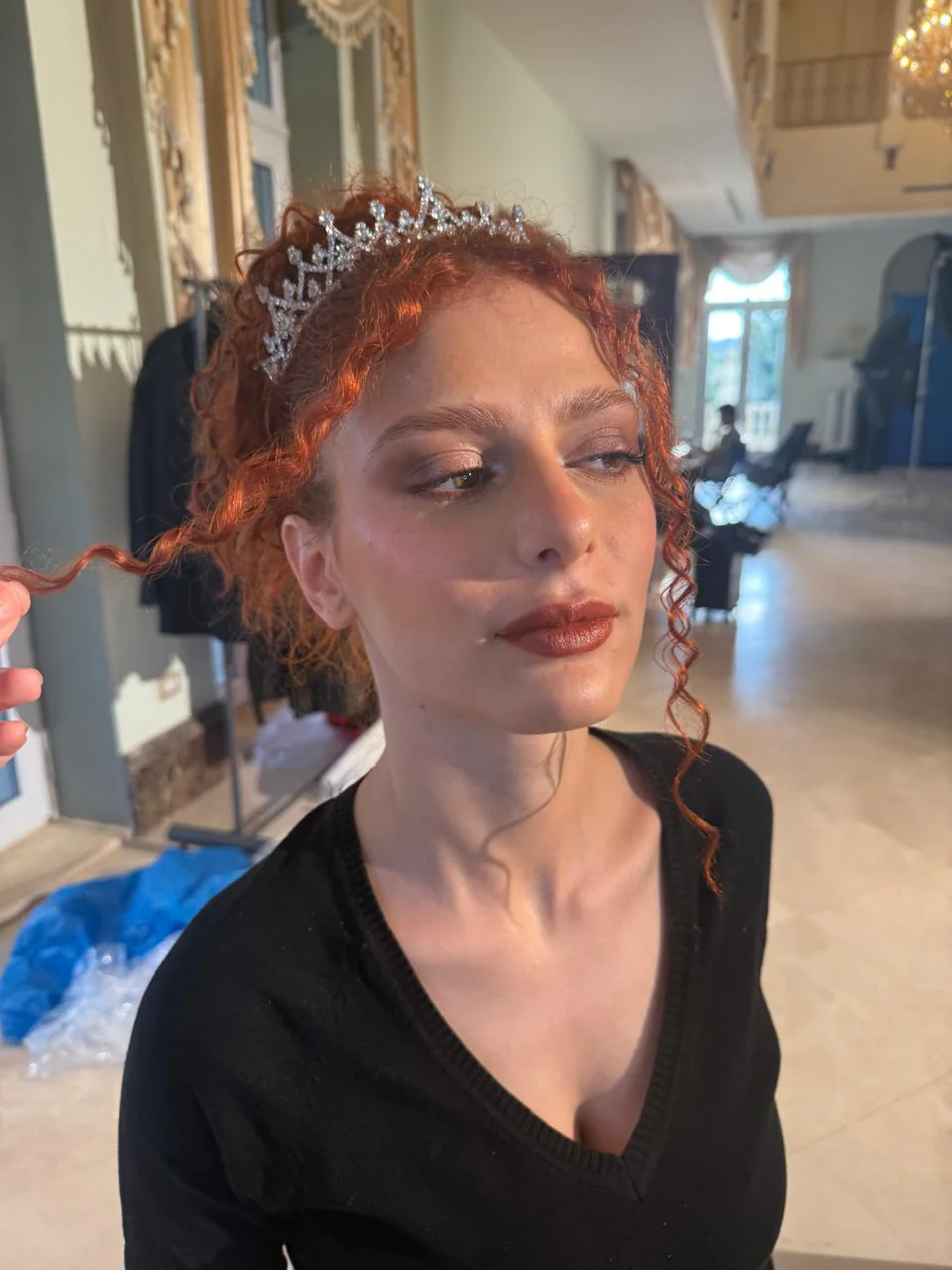 Close three-quarter performer portrait in an interior: curly red hair with tiara, warm brown/bronze eye color, defined lashes, red-brown lips and black top; a hand holds a curl at left.