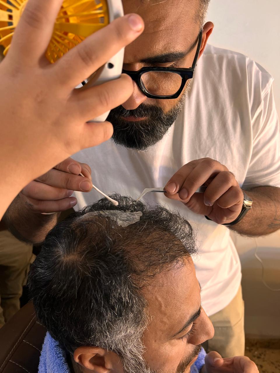 Close BTS photo of a person using a cotton swab and tweezers at a seated performer's hair; a small fan and another hand partly obscure the upper scene.