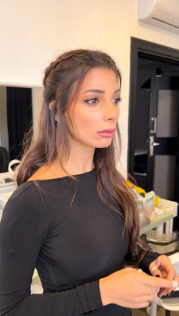 Three-quarter performer portrait in a makeup-room setting: long brown hair with a braided section, dark winged eyeliner/defined lashes, warm cheeks and pink-brown lips; black top and hands visible. Mirror/table products in the background.