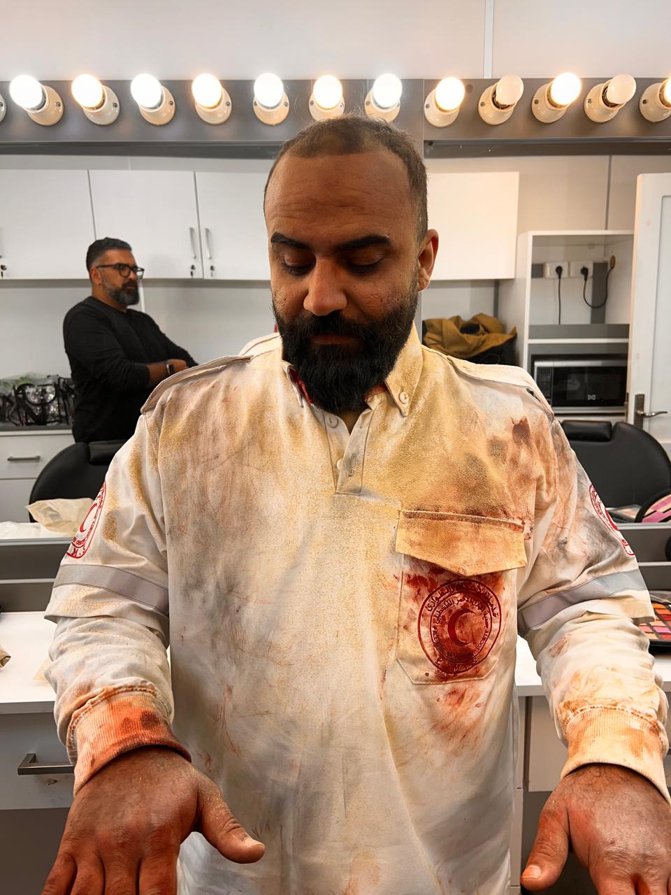 Bearded performer in a dressing-room chair wearing a pale paramedic-style uniform with extensive red/brown costume staining; forehead and hands show dirt-like tonal detail. Mirrors and bulbs behind.