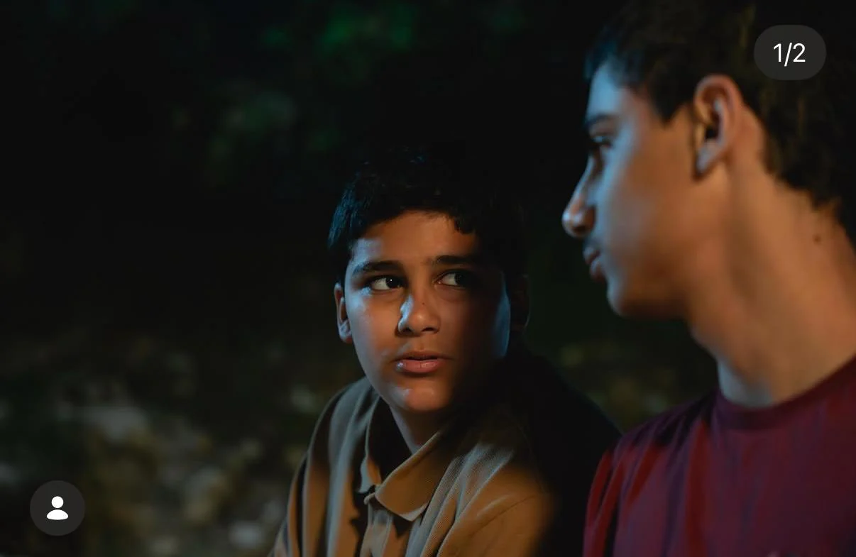 Landscape night scene of two young male performers speaking; Instagram carousel 1/2 badge and tagged-person icon are embedded. No printed project title.